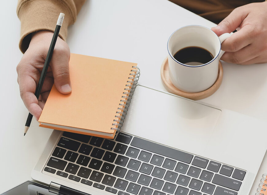 Hands holding spiral notebook, pencil, coffee, laptop