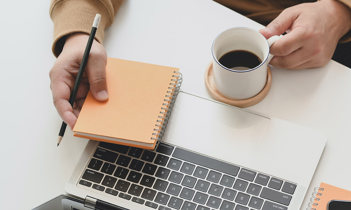 Hands holding spiral notebook, pencil, coffee, laptop