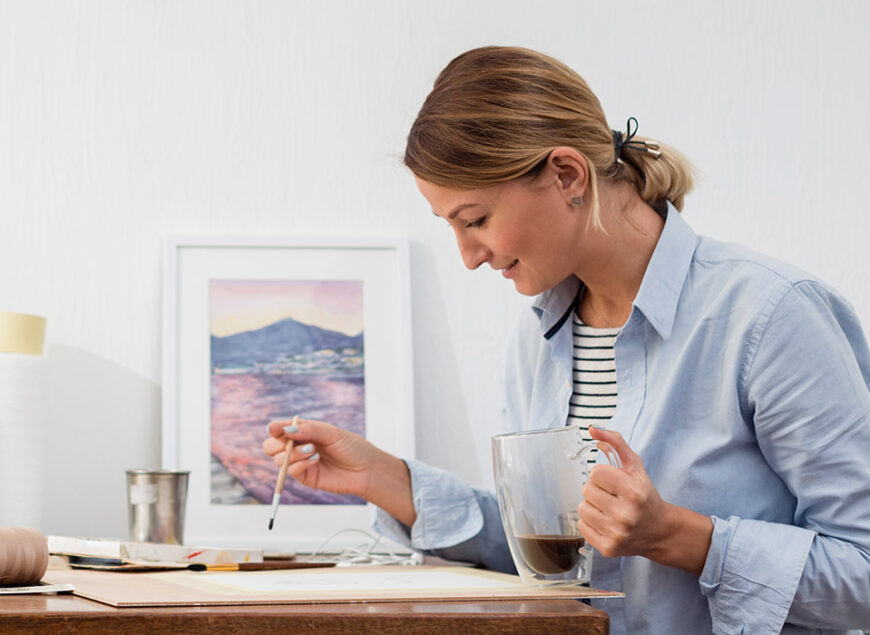 Artist painting at desk while holding coffee